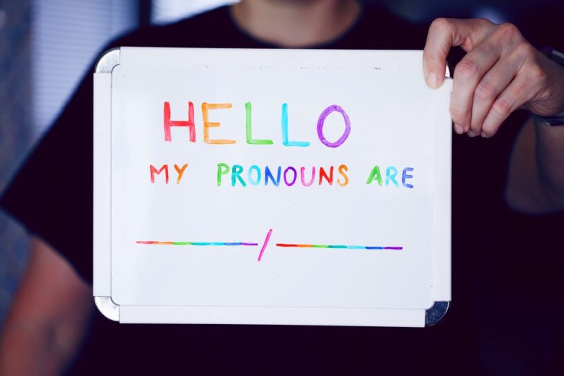 Ein Mensch hält ein Schild hoch. In Regenbogenschrift steht darauf: Hello My Pronouns Are